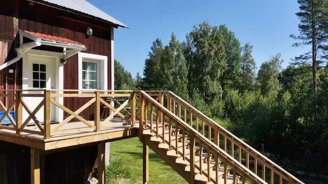 Rood houten huis met een houten trap en balustrade, omgeven door groene bomen en een blauwe lucht.