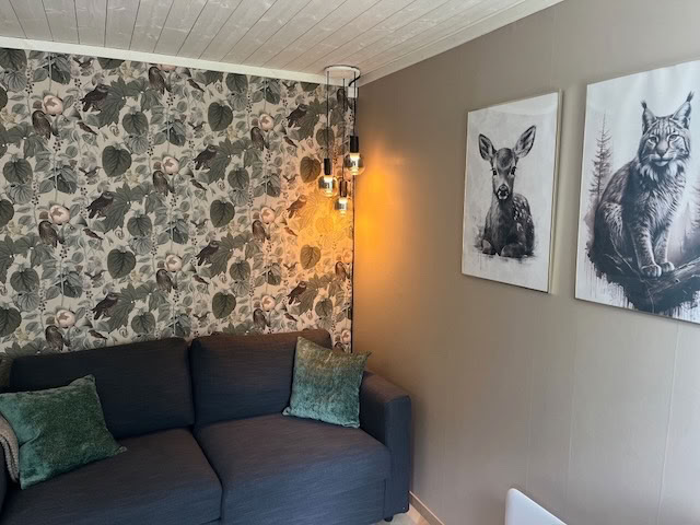 Donkergrijze bank met groene kussens, bloemenbehang op de muur, twee zwart-wit dierenprints aan de muur en een hanglamp met meerdere gloeilampen.